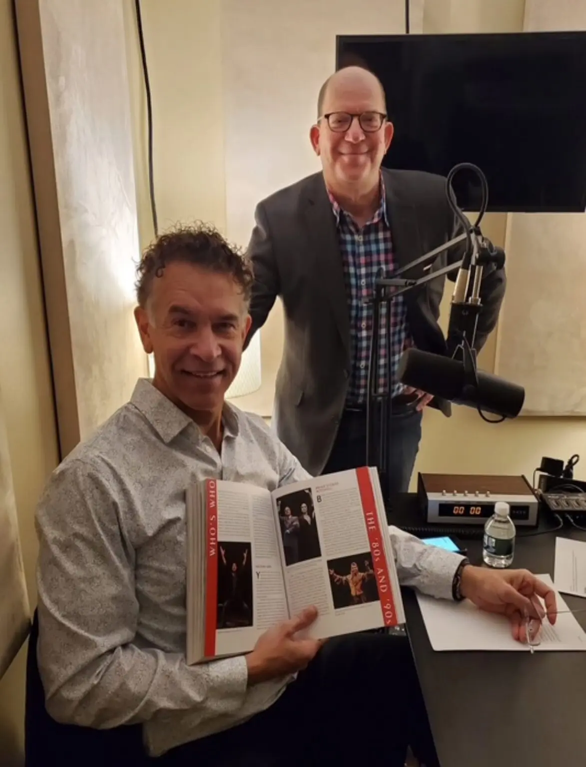 Two men in a recording studio, one holding a book.